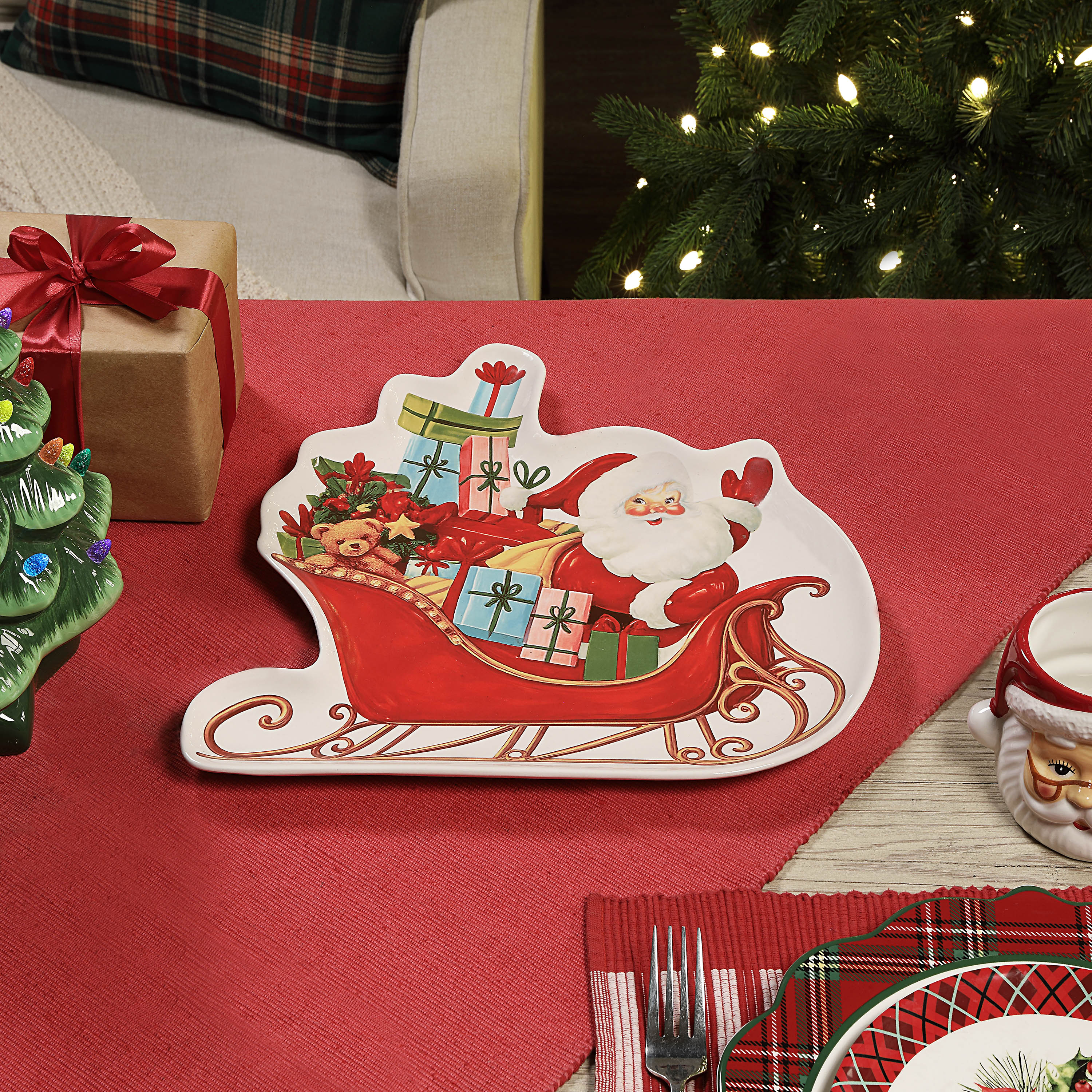 A Christmas-themed ceramic plate shaped like Santa's sleigh, featuring an illustration of Santa Claus carrying wrapped gifts, displayed on a red table runner with holiday decorations in the background.