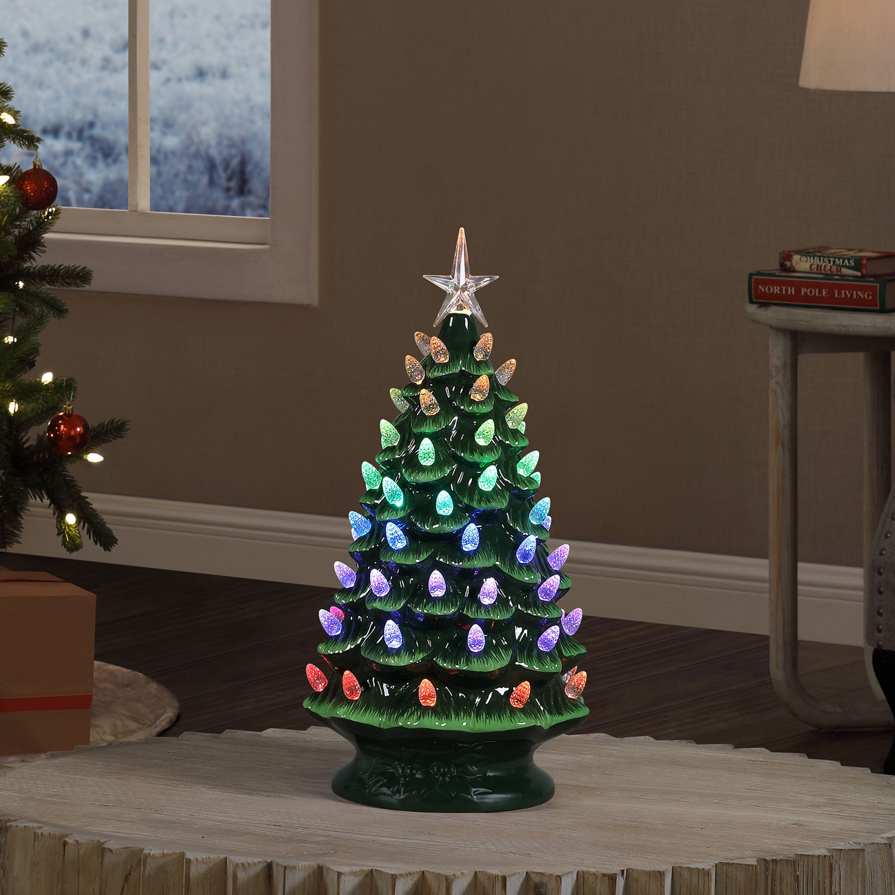 A small green ceramic Christmas tree with multicolored lit lights and a star topper, displayed on a round table in a cozy indoor room setting.
