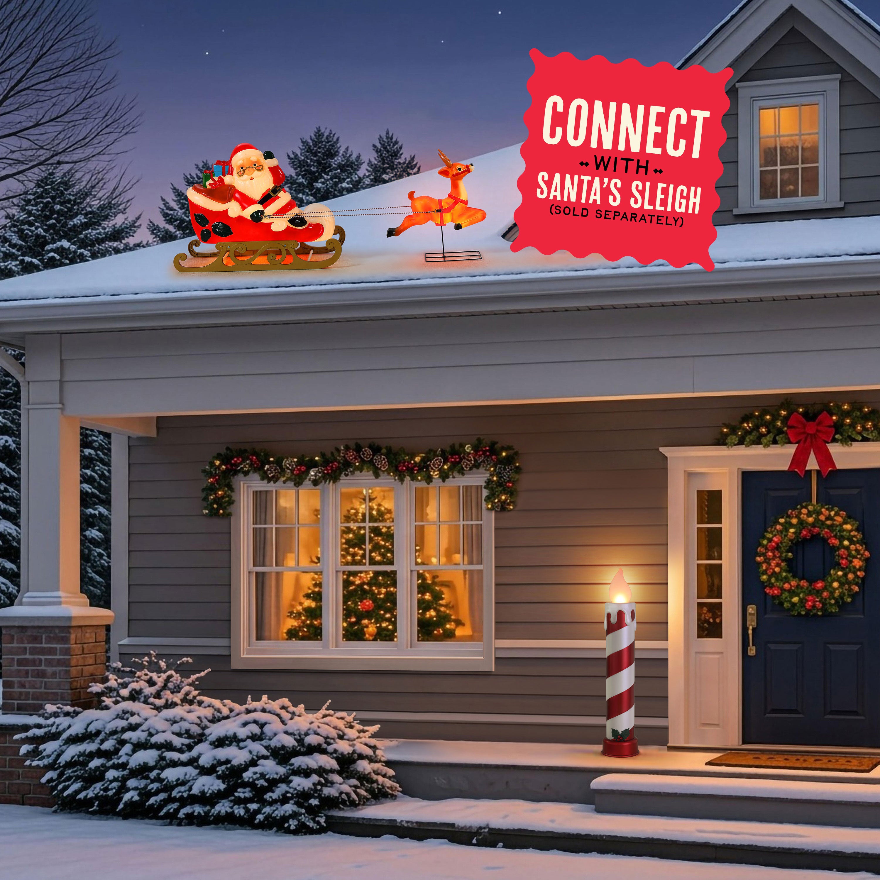Christmas outdoor holiday decorations on a snow-covered house roof, featuring an illuminated Santa in a sleigh and a reindeer, with a red promotional badge that says 'CONNECT WITH SANTA'S SLEIGH (SOLD SEPARATELY)'. The house is decorated with garlands, a wreath, a lit candle post, and warm windows glowing in the evening.
