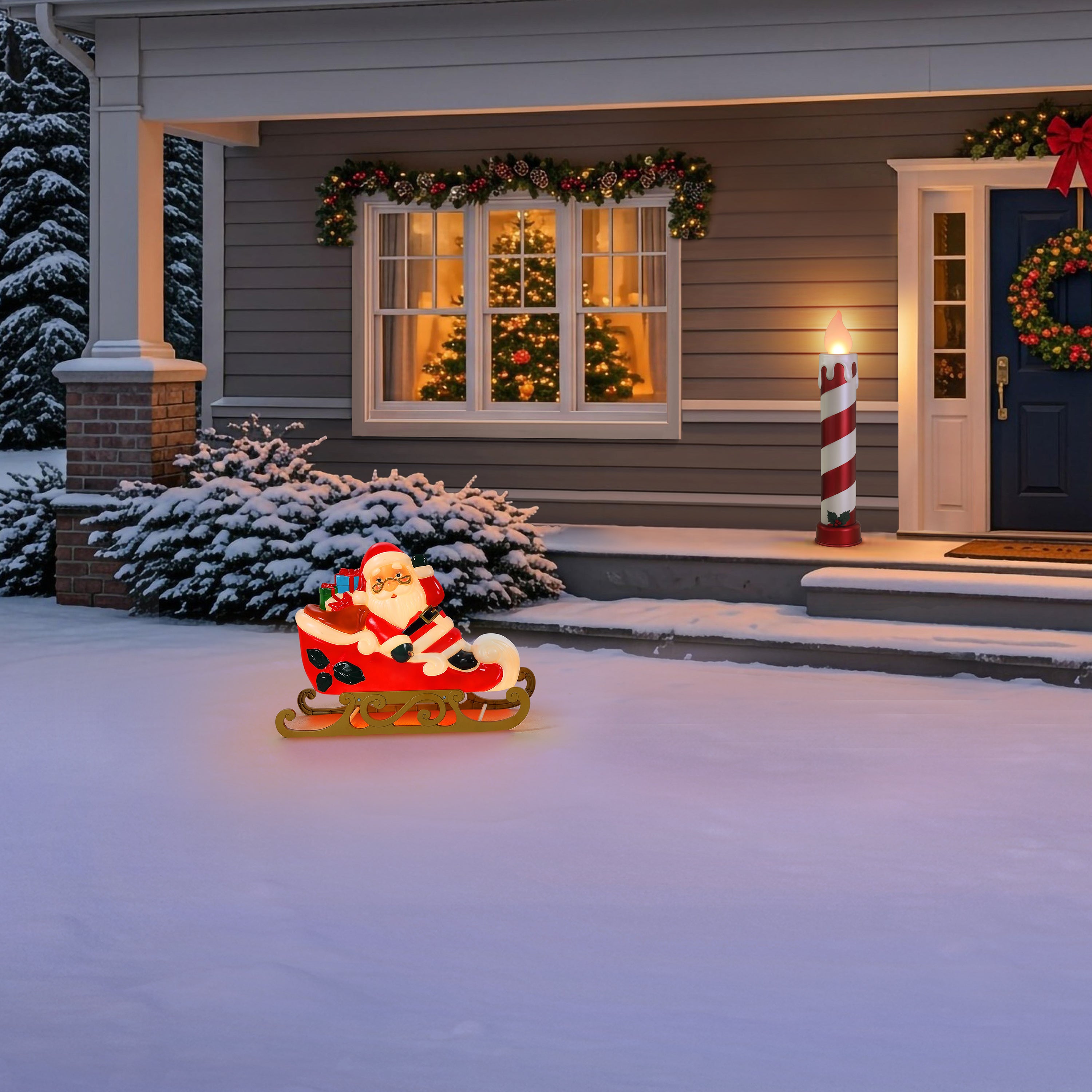 A Santa Claus Christmas light in a red sleigh sits on the snowy front yard in front of a decorated house with a lit window, wreath, and candy cane.