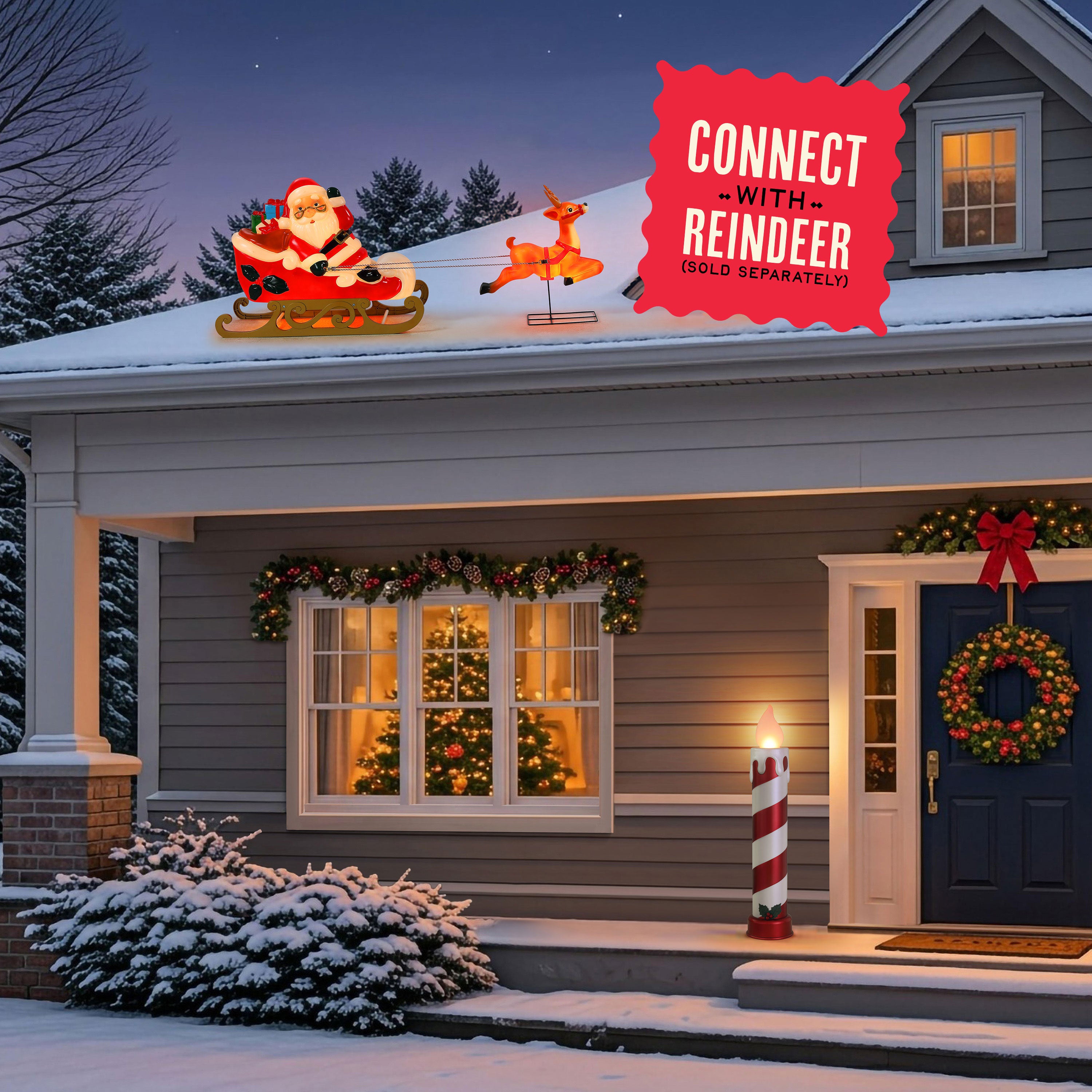 Christmas outdoor holiday decorations on a snow-covered house roof, featuring a glowing Santa Claus in a sled and a reindeer, with a red promotional badge that reads 'CONNECT WITH REINDEER (GOLD SEPARATELY)'. The house is decorated with garlands, a wreath, a lit candle post, and warm windows in the evening.