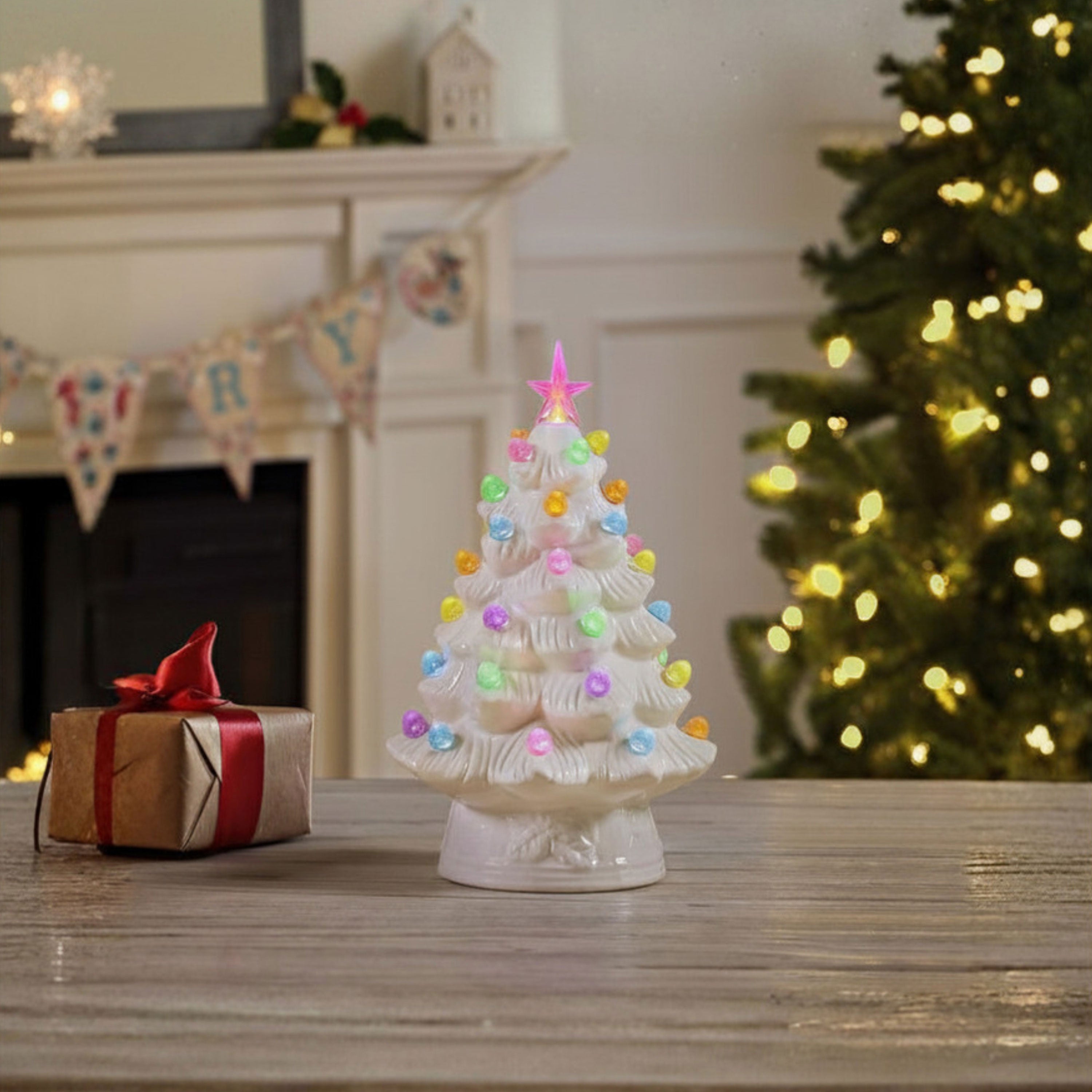 A small white ceramic Christmas tree ornament with colorful glowing lights and a pink star topper, displayed on a wooden table in a cozy holiday room setting with a fireplace, gift box, and a blurred lit Christmas tree in the background.