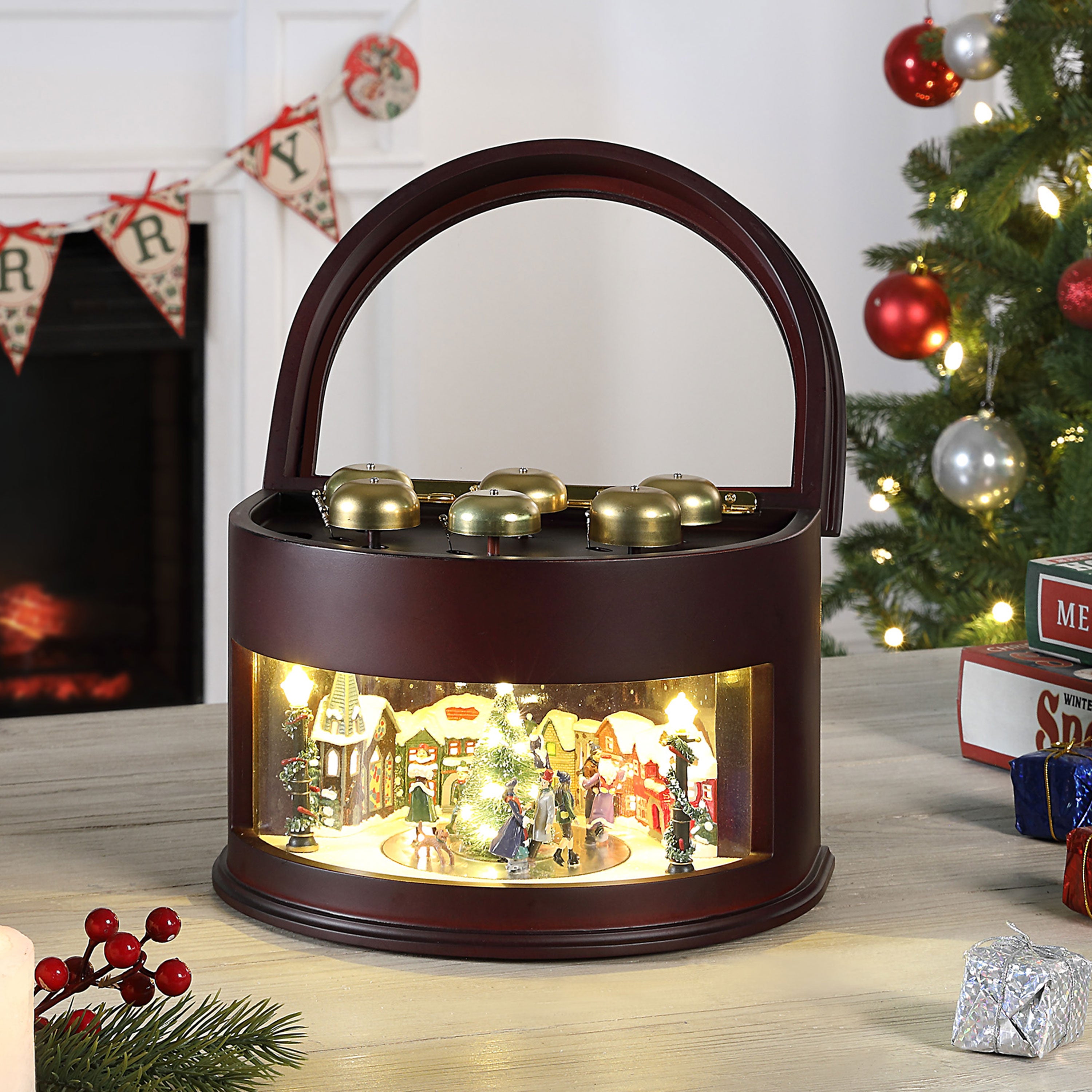 A round wooden music box with a handle and a lit Christmas nativity scene inside, placed on a tabletop next to a decorated Christmas tree and fireplace.