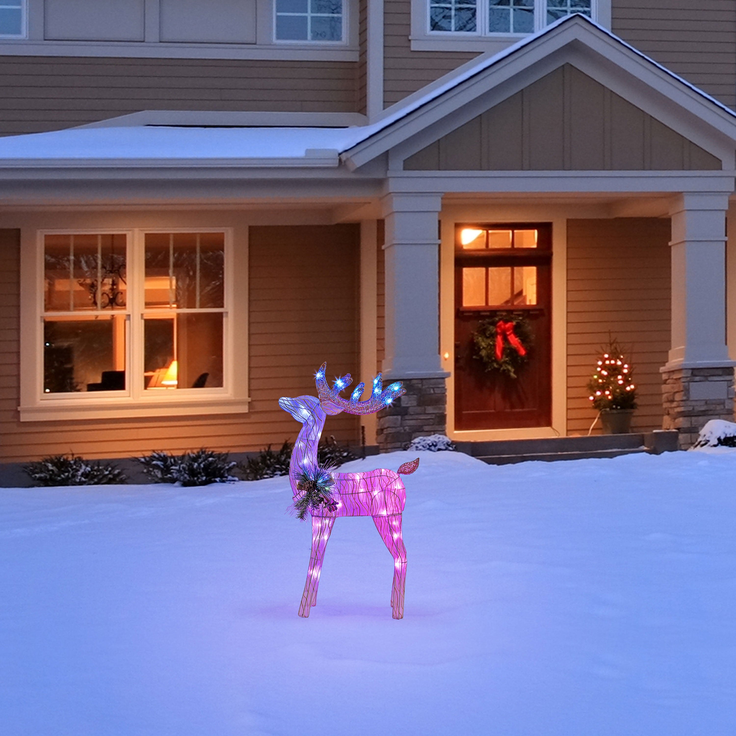 lose-up of Mr. Christmas 3 ft. Animated Fiber-Optic Reindeer glowing in the snow on a front yard, holiday-lit house in background