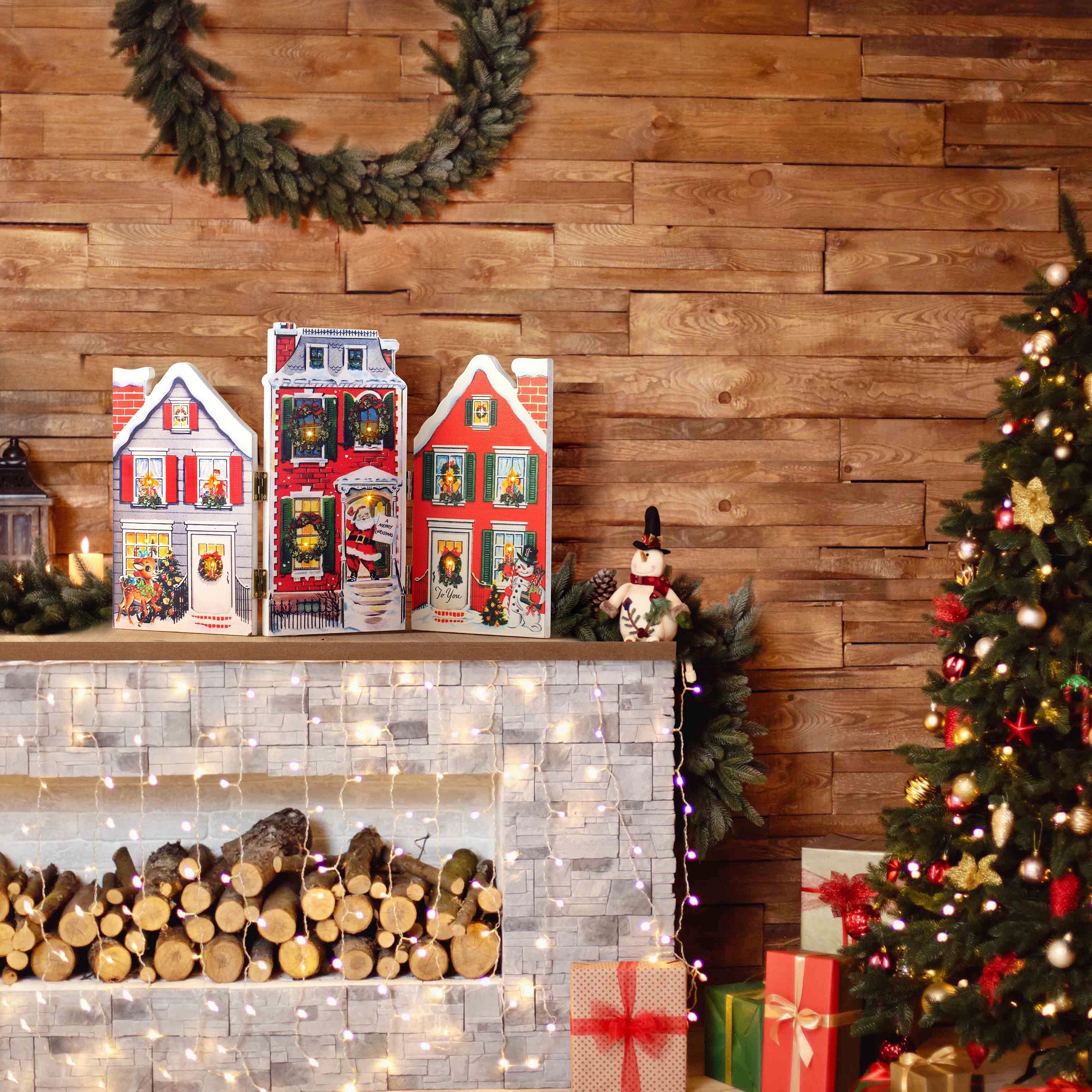 Mr. Christmas Folding Village Scene displayed on a decorated fireplace mantel near a Christmas tree, glowing with warm white LED lights.