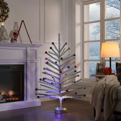 A silver tinsel Christmas tree with colorful ornaments stands in a cozy living room near a window and fireplace, lit with multicolor LED lights.