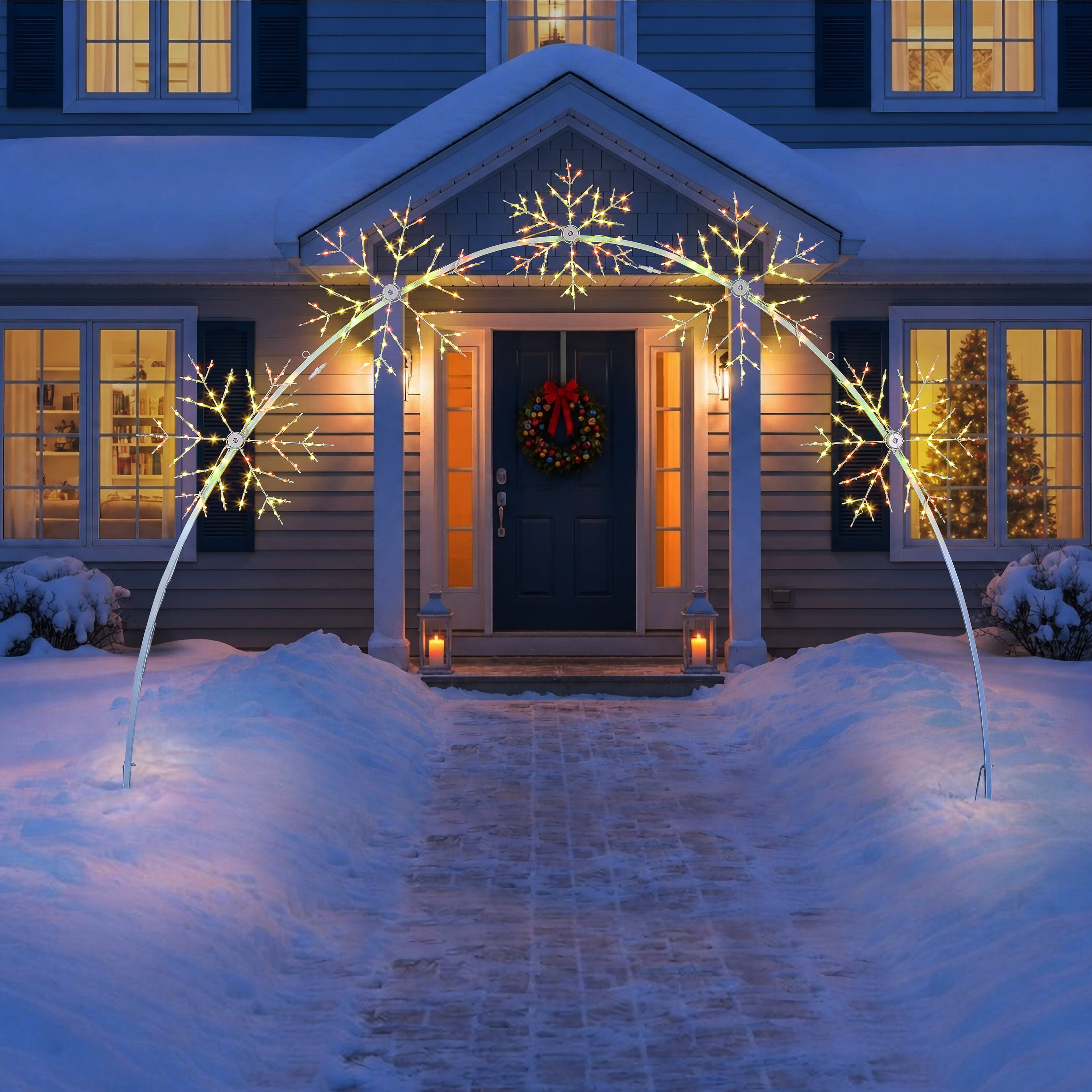 Snow-covered front yard with a white arched holiday light fixture shaped like snowflakes, illuminated in warm white light above a house entrance with a wreath and lit windows.
