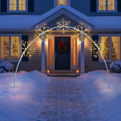 Snow-covered front yard with a white arched holiday light fixture shaped like snowflakes, illuminated in warm white light above a house entrance with a wreath and lit windows.