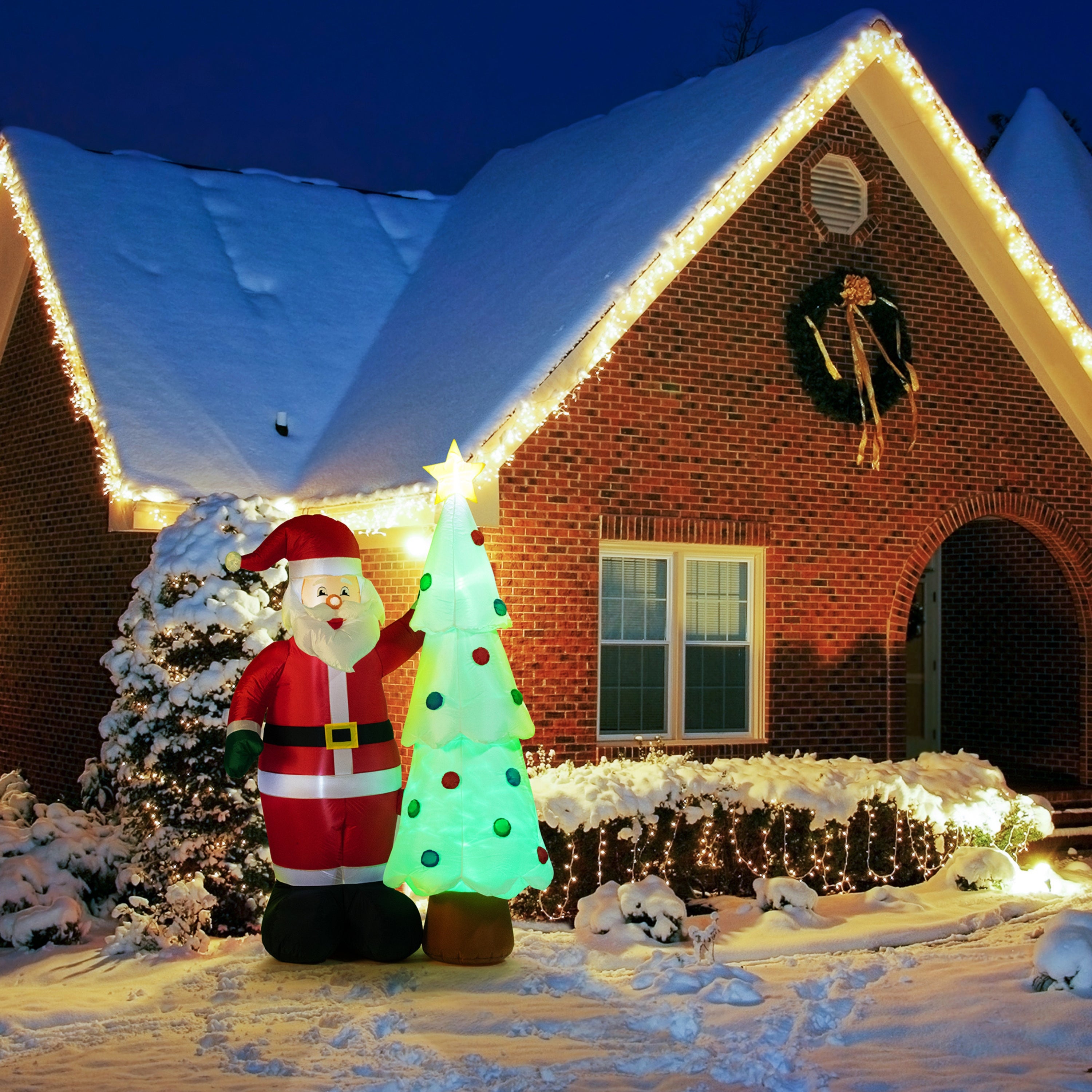 A snow-covered house with Christmas lights and a large wreath displays the 7 ft. Outdoor Lit Inflatable - Santa and Tree, made from weather-resistant materials, glowing brightly in the front yard at night.