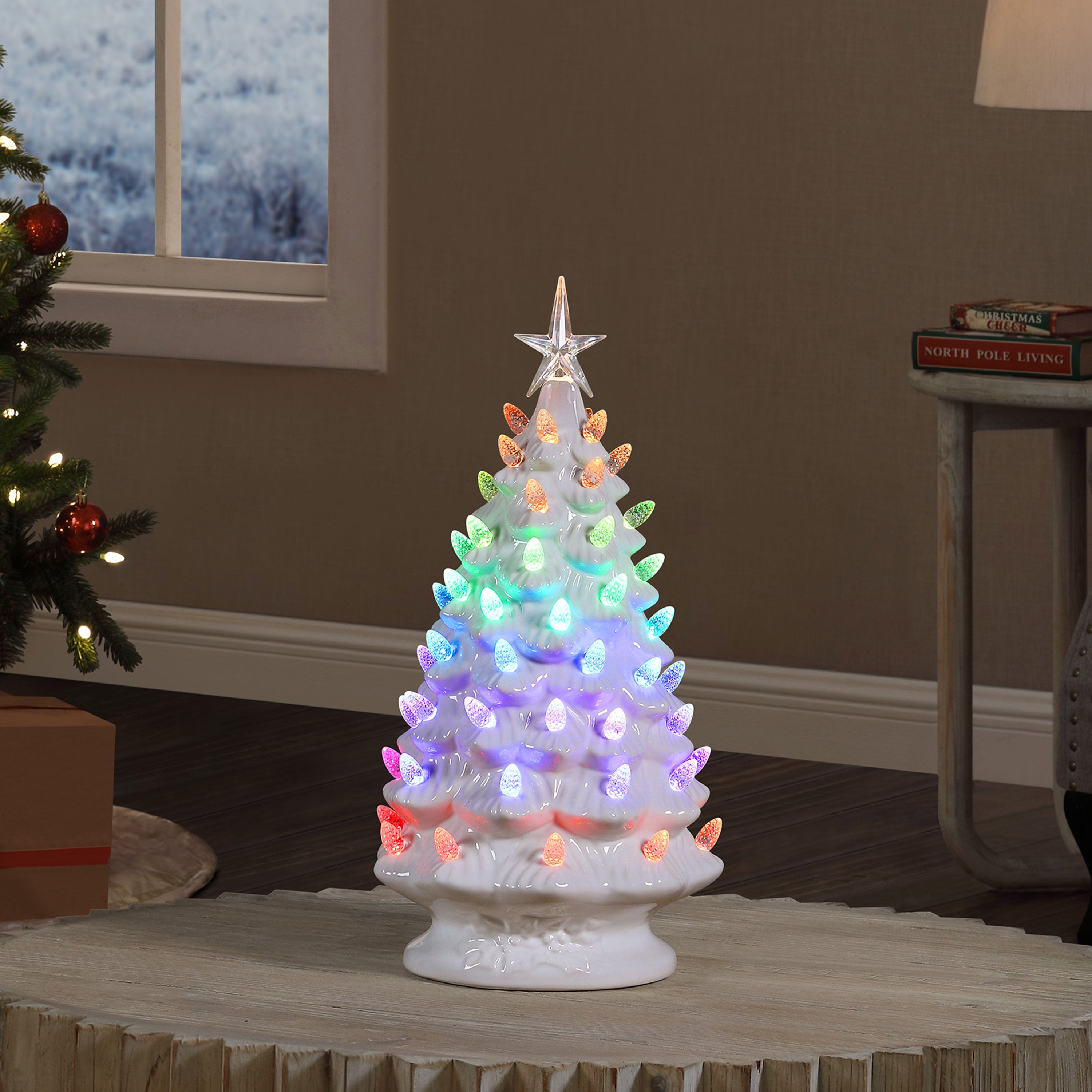 A small white ceramic Christmas tree with multicolor LED lights glowing from the tips, topped with a clear star, displayed on a table in a cozy room setting.