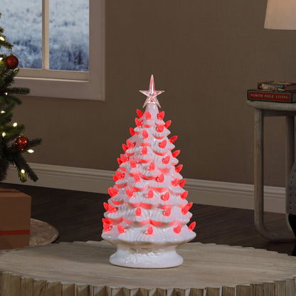 A white ceramic Christmas tree with glowing pink LED lights and a pink star topper, displayed on a table in a cozy living room setting.