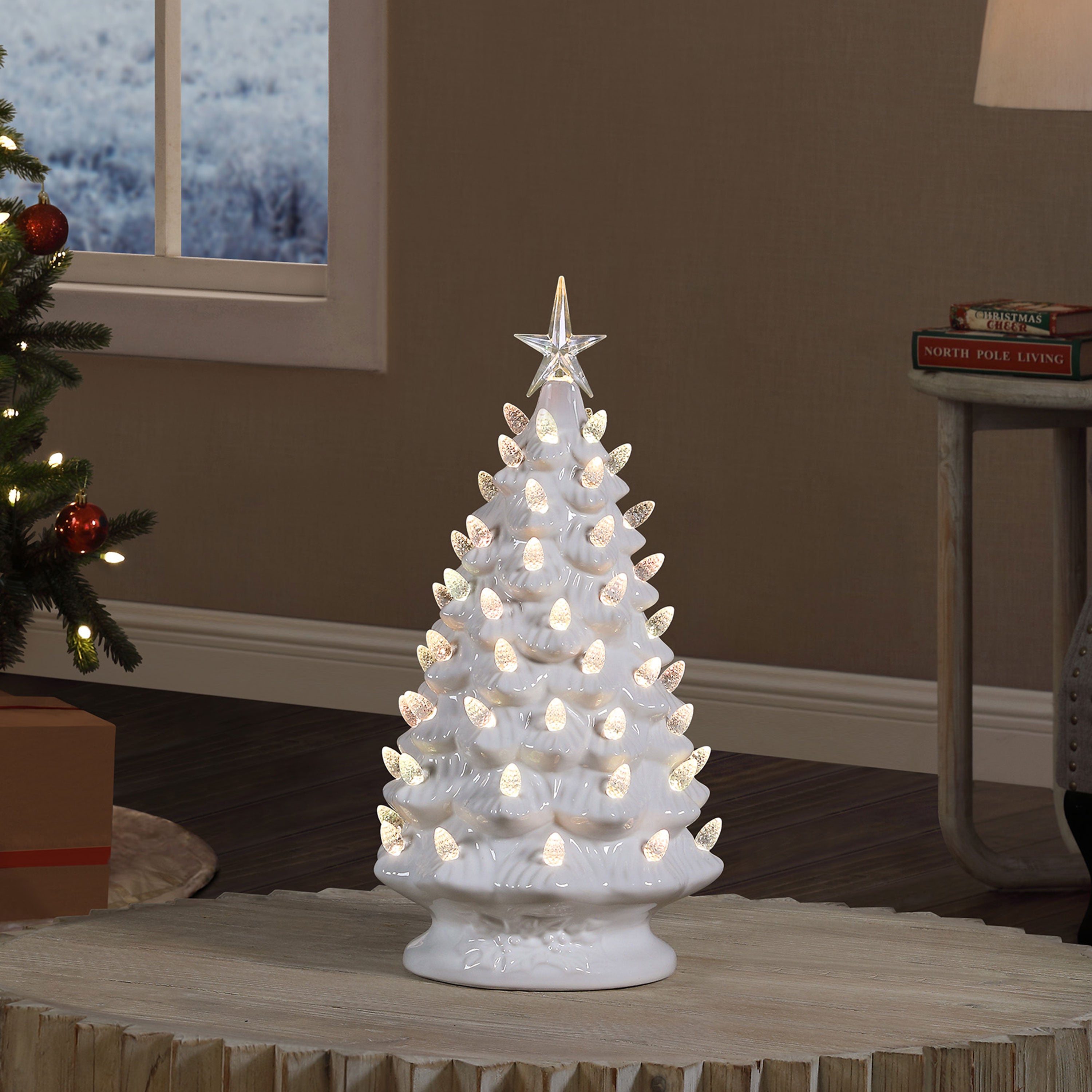 A white ceramic Christmas tree table lamp with warm white LED lights glowing inside, topped with a clear star, displayed on a wooden table in a cozy living room setting.