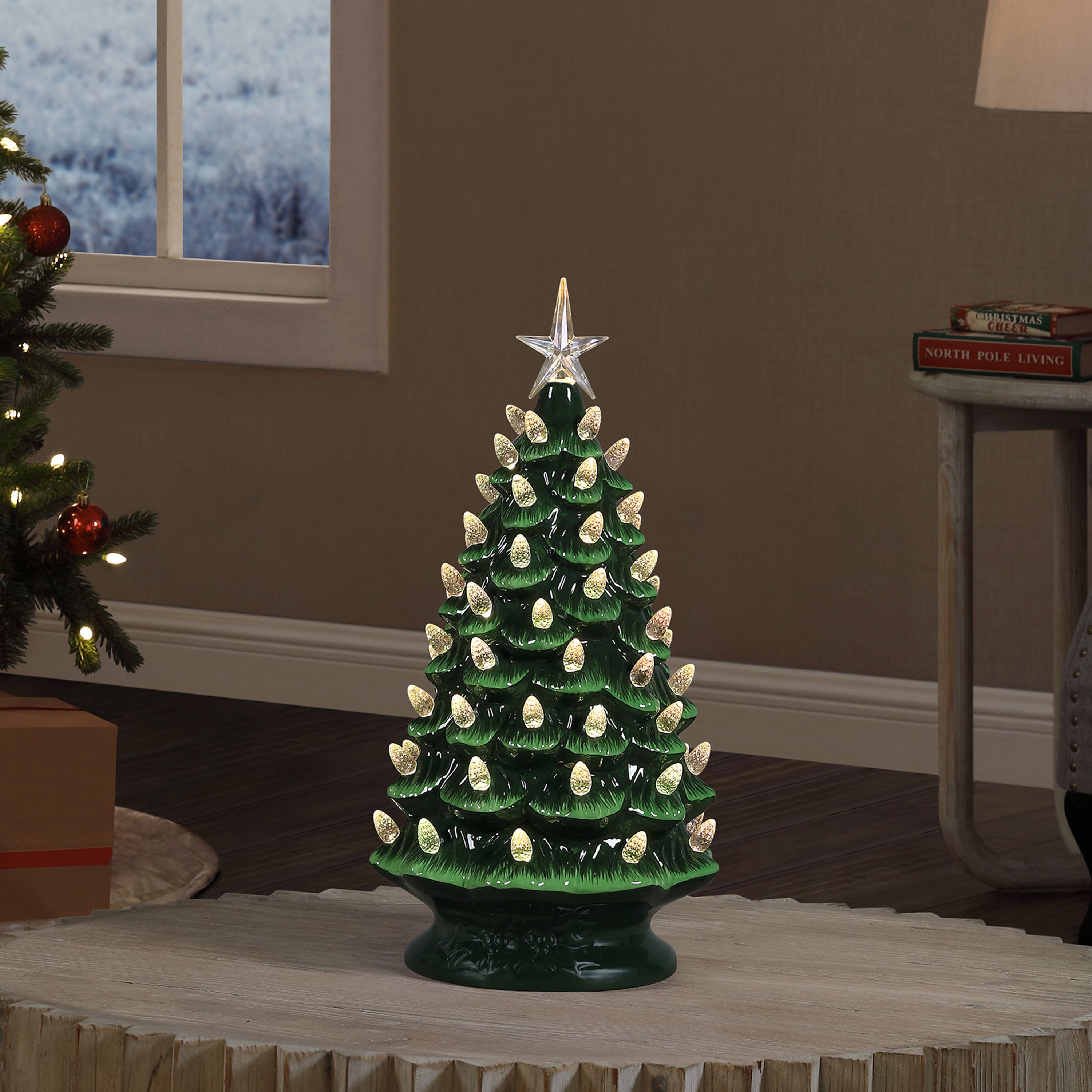 A green ceramic Christmas tree with lit white bulbs and a gold star topper, displayed on a round table in a living room setting.