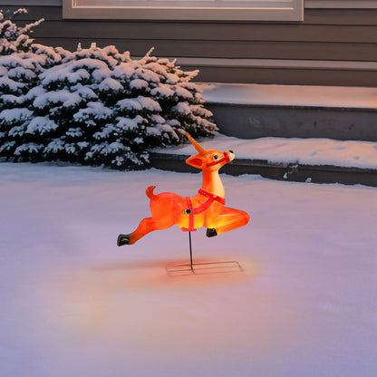 An illuminated orange-red reindeer yard light on a metal stand, photographed outdoors in snow with a house and snow-covered shrub in the background.