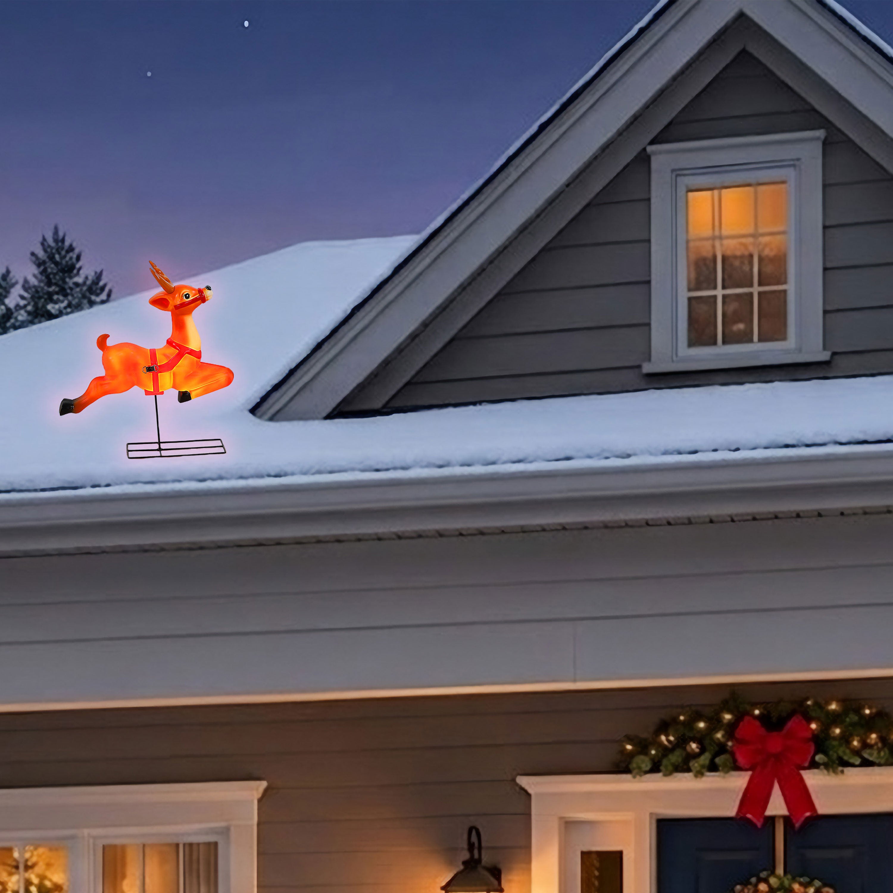 An illuminated orange Christmas flying reindeer light mounted on a snow-covered house roof at dusk, with the house exterior, lit windows, and holiday wreath visible below.
