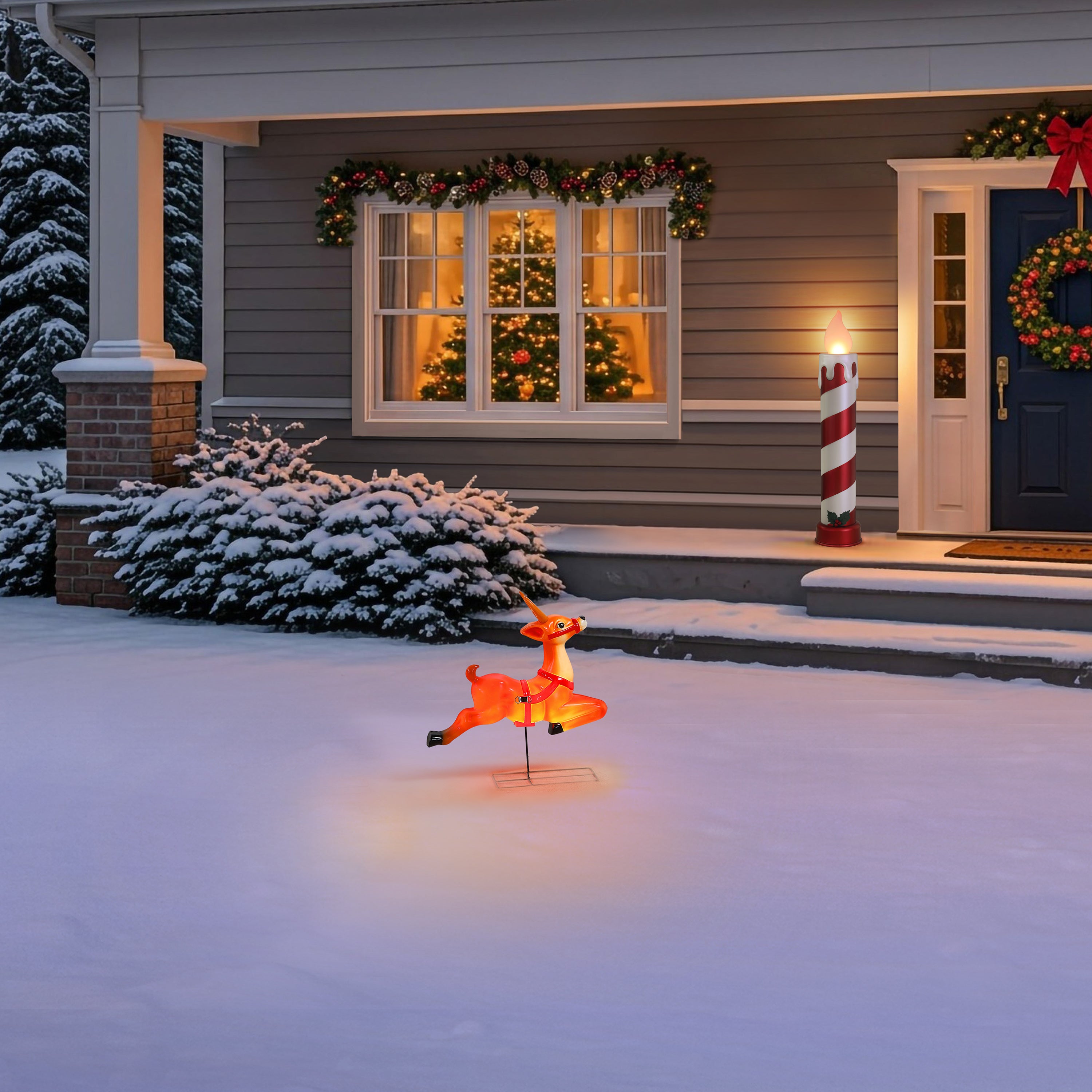A lit orange Christmas flying reindeer yard light stands on snow in front of a decorated house porch at dusk.