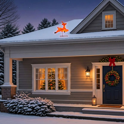 A snow-covered suburban house at dusk with holiday decorations: a glowing orange reindeer light on the roof, a wreath and red bow on the front door, lanterns on the porch, and warm lights inside the windows.