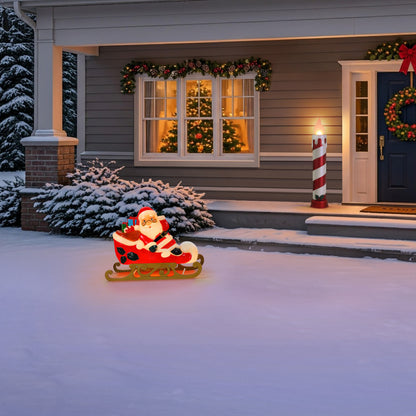A Santa Claus Christmas light in a red sleigh sits on the snowy front yard in front of a decorated house with a lit window, wreath, and candy cane.