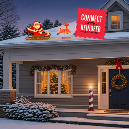 Christmas outdoor holiday decorations on a snow-covered house roof, featuring a glowing Santa Claus in a sled and a reindeer, with a red promotional badge that reads 'CONNECT WITH REINDEER (GOLD SEPARATELY)'. The house is decorated with garlands, a wreath, a lit candle post, and warm windows in the evening.