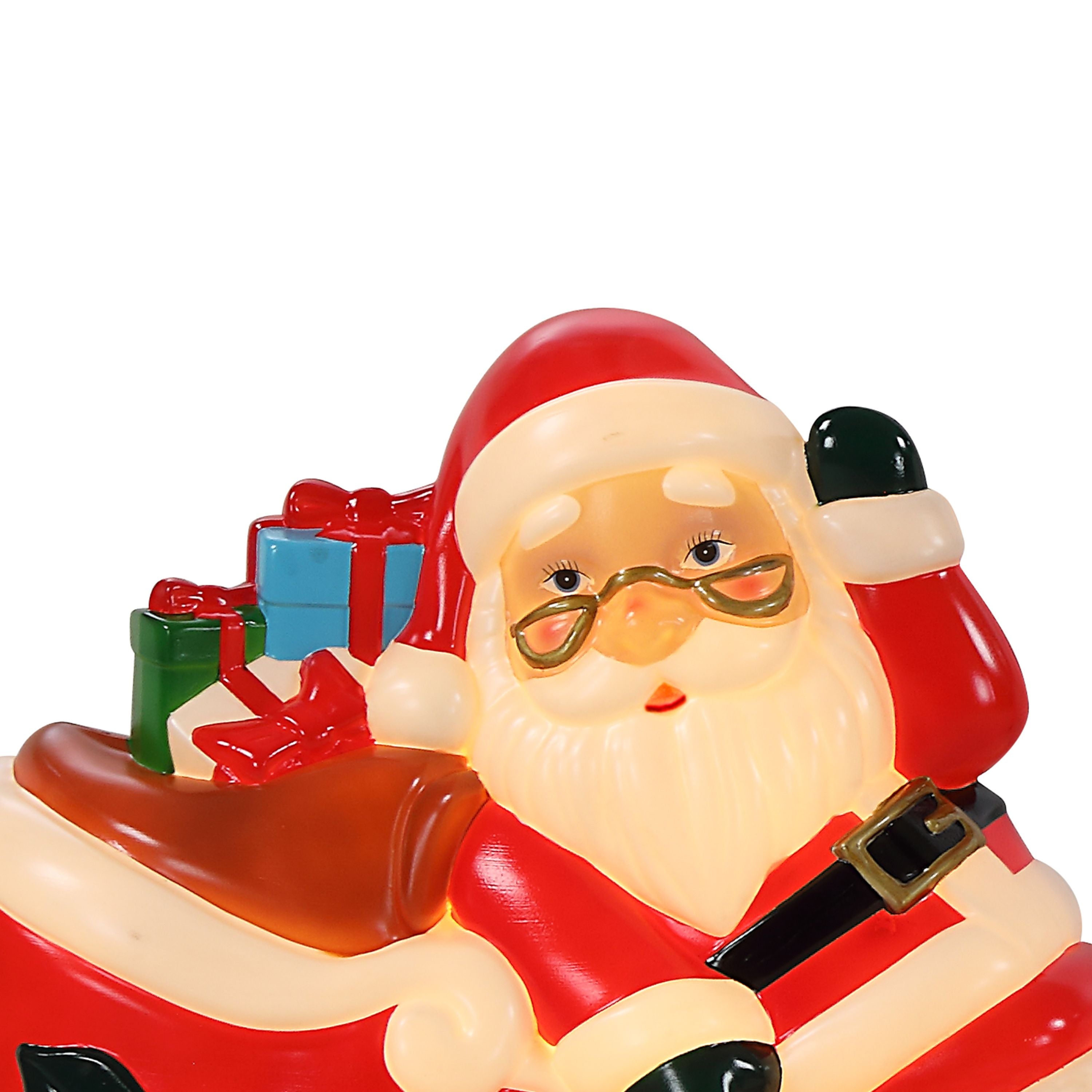 A close-up of a Santa Claus figurine or decorative object seated in a sleigh, with red and white clothing, gold glasses, a black belt, and small gift boxes on his back, shown against a plain white background.
