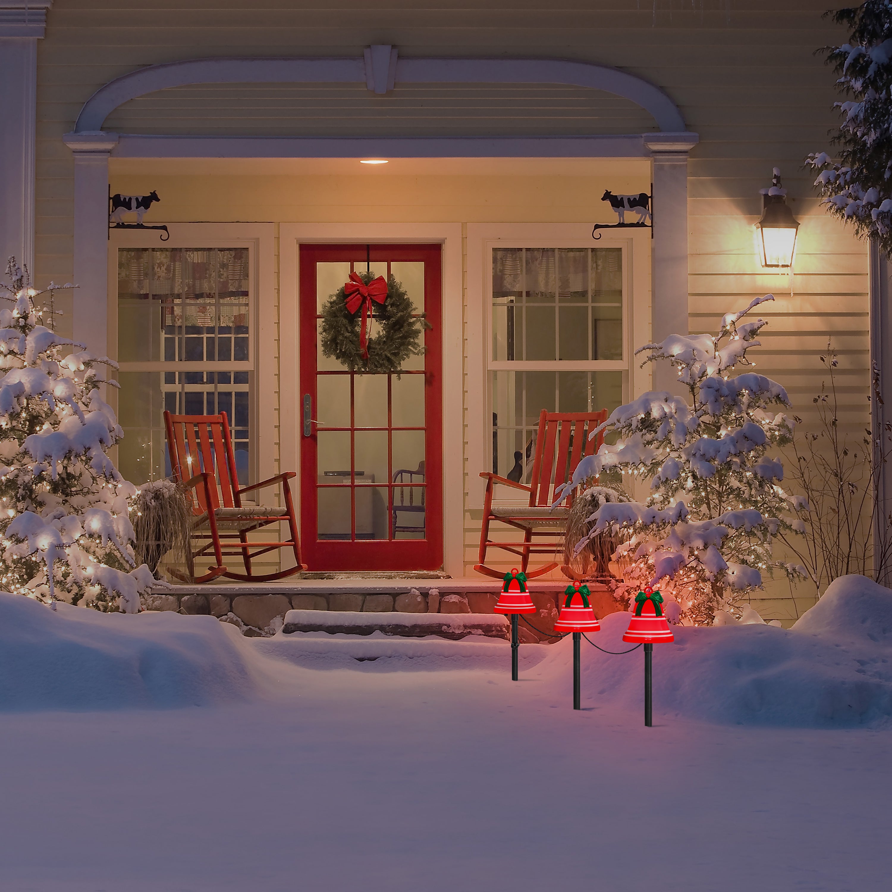 A snowy front porch at night with a red door, white rocking chairs, decorated Christmas trees, and red bell-shaped outdoor lights on stakes in the snow.