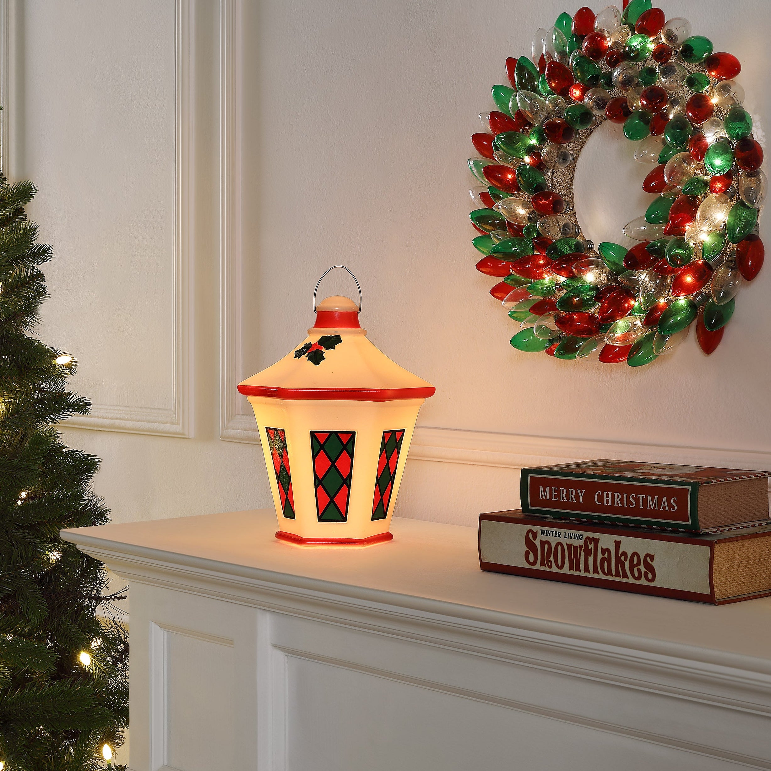 A lit Christmas table lantern lamp with a cream body, red trim, and red-and-black diamond pattern, placed on a white mantel beside a wreath, books, and a Christmas tree.