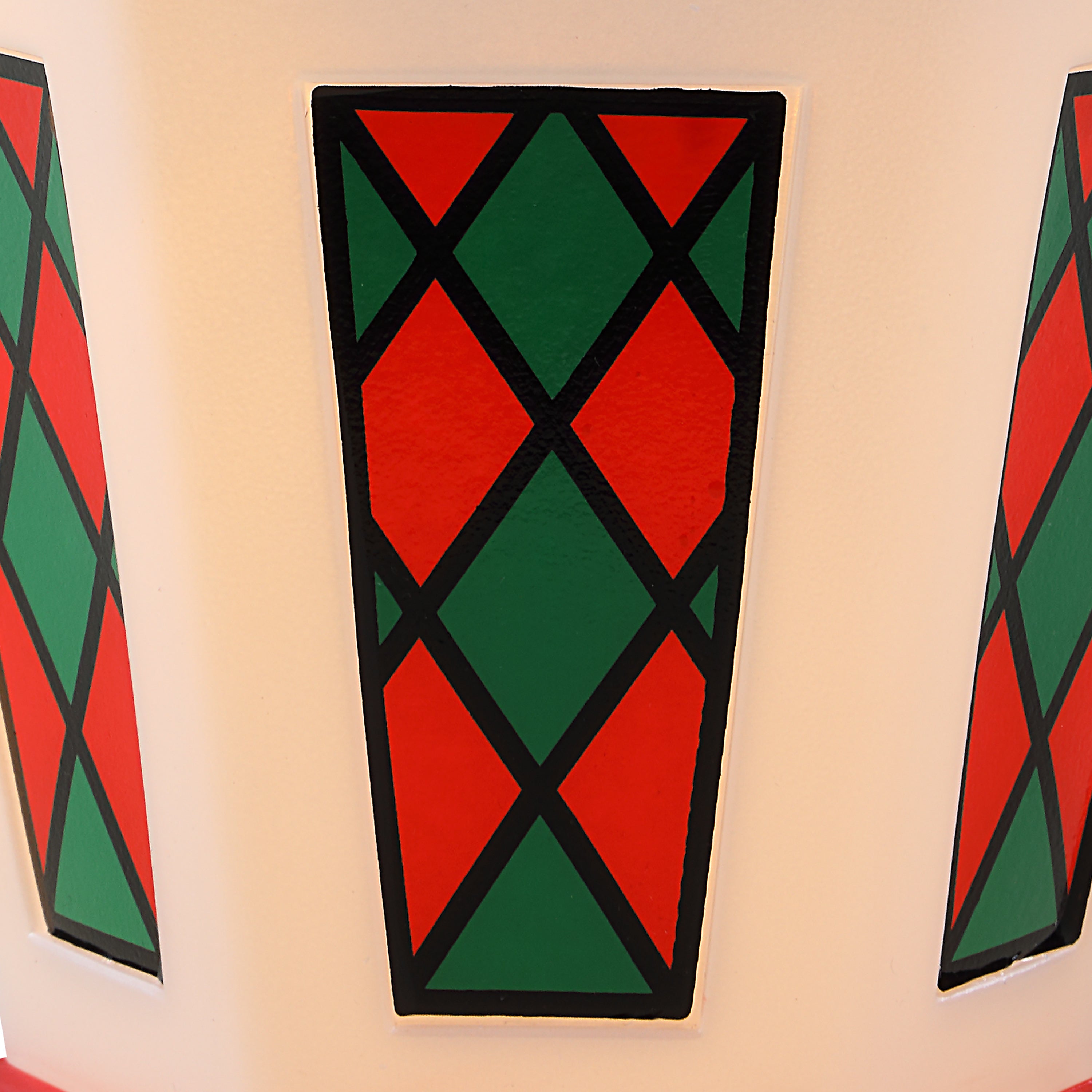 A close-up of a decorative geometric pattern on a Christmas lantern featuring alternating red and green diamond panels outlined in black, mounted on a light beige wall.
