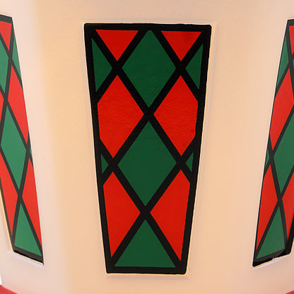 A close-up of a decorative geometric pattern on a Christmas lantern featuring alternating red and green diamond panels outlined in black, mounted on a light beige wall.