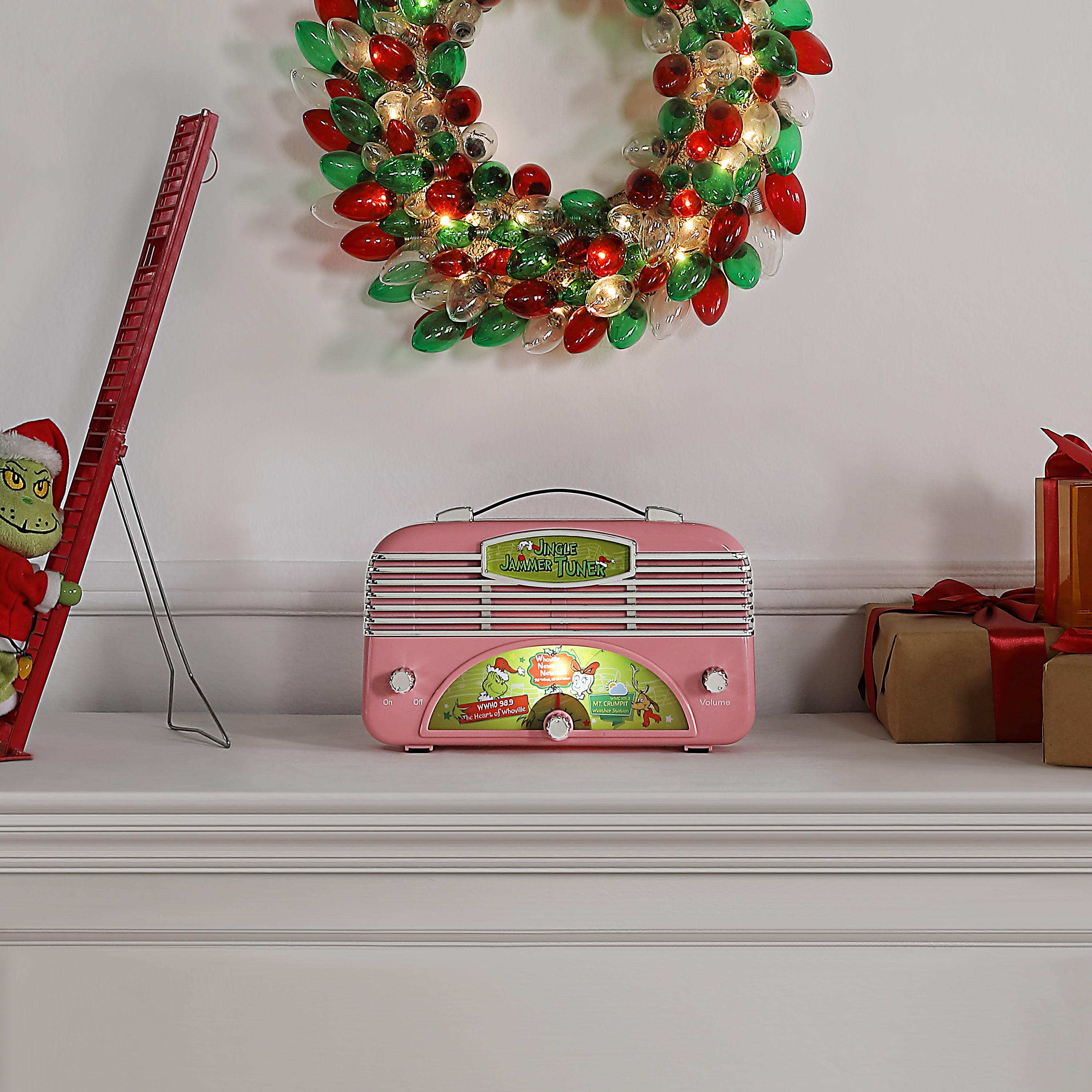 A pink grinch retro-style radio with a Christmas train illustration on the front, displayed on a white mantelpiece below a holiday light wreath. The radio has silver vents, white knobs, and a small round dial.