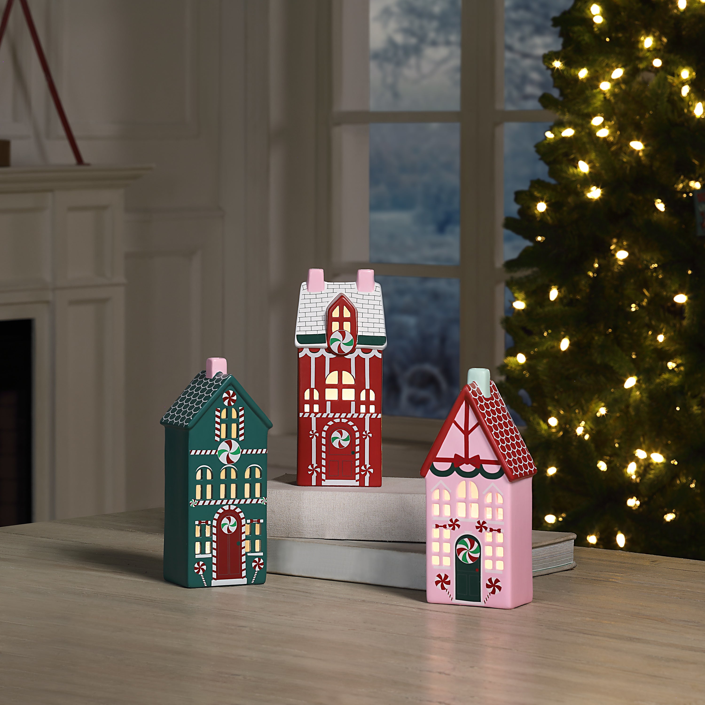 Three small decorative flat Christmas house ornaments in teal, red, and pink, displayed on stacked books on a wooden table with a Christmas tree and fireplace in the background.
