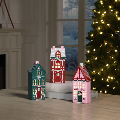 Three small decorative flat Christmas house ornaments in teal, red, and pink, displayed on stacked books on a wooden table with a Christmas tree and fireplace in the background.