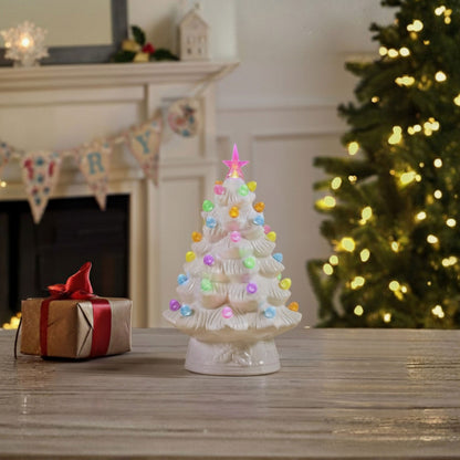 A small white ceramic Christmas tree ornament with colorful glowing lights and a pink star topper, displayed on a wooden table in a cozy holiday room setting with a fireplace, gift box, and a blurred lit Christmas tree in the background.