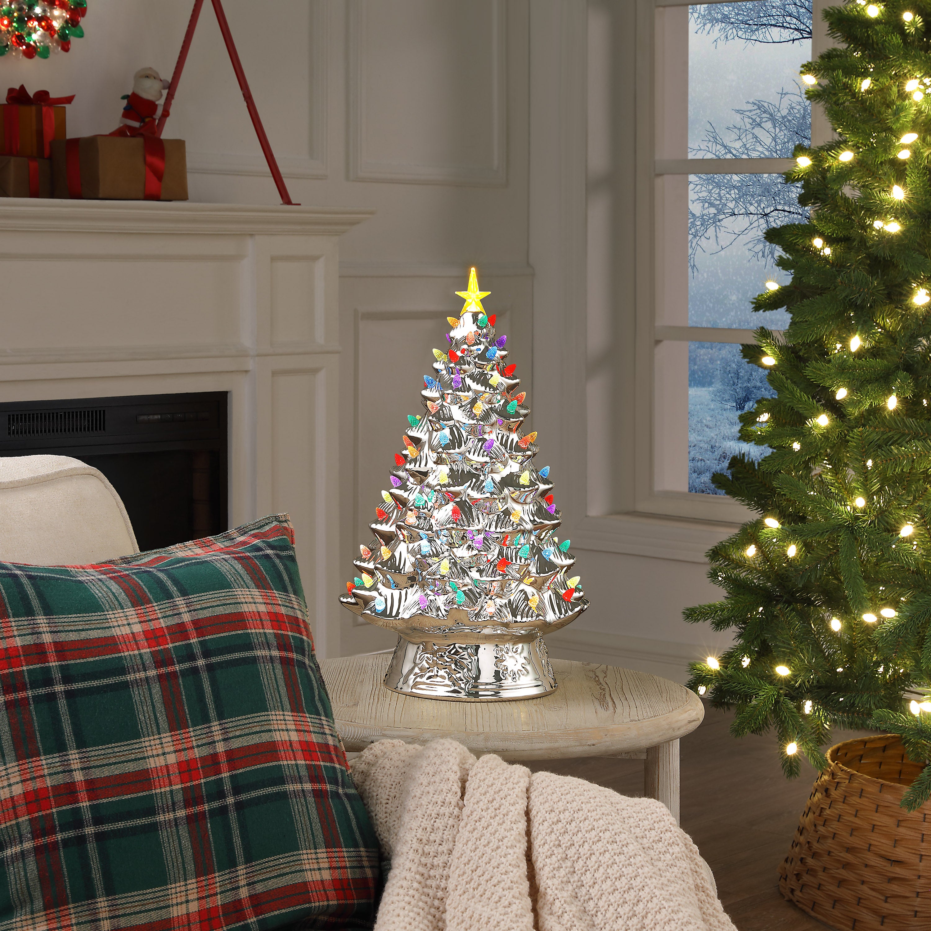 A silver Christmas tree with colorful ornaments and a gold star sits on a small table in a cozy living room setting next to a larger lit Christmas tree.