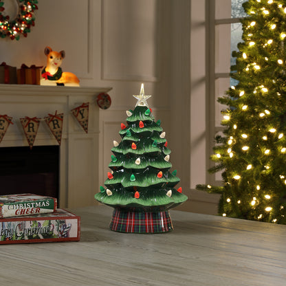 A vintage-style ceramic Christmas tree centerpiece with a green tiered body, red, white, and green ornament accents, a white star topper, and a plaid base, displayed on a tabletop in a cozy holiday room setting.