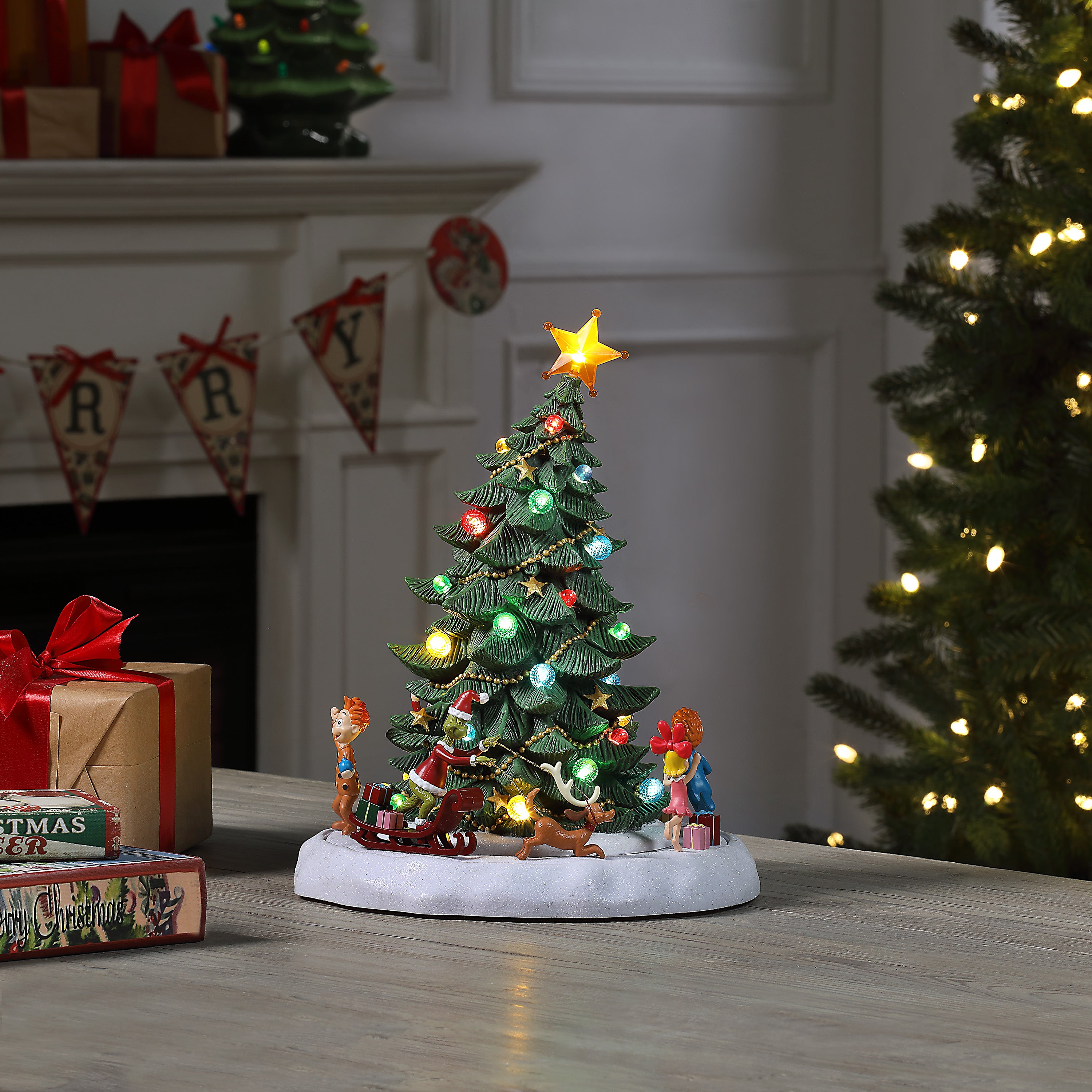 A small lit Christmas tree figurine with colorful ornaments, a star topper, and holiday characters on a white base, displayed on a tabletop in a decorated holiday room with wrapped gifts and a larger tree in the background.