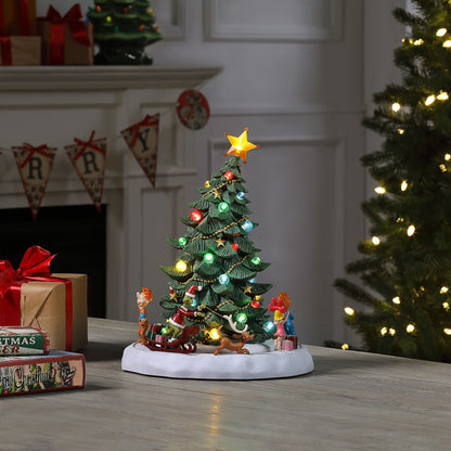 A small lit Christmas tree figurine with colorful ornaments, a star topper, and holiday characters on a white base, displayed on a tabletop in a decorated holiday room with wrapped gifts and a larger tree in the background.