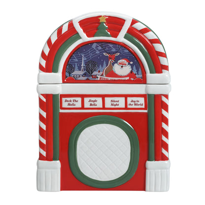 A Christmas-themed jukebox-shaped container or ornament with red, white, and green candy cane stripes, a festive arched top, and a winter scene panel showing Santa and a reindeer above four song labels: 'Deck The Halls,' 'Jingle Bells,' 'Silent Night,' and 'Joy to the World.'.
