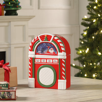 A red and white candy cane-themed jukebox-shaped Christmas ornament or decorative container sits on a tabletop in a holiday living room setting, with a lit Christmas tree and wrapped gifts in the background.