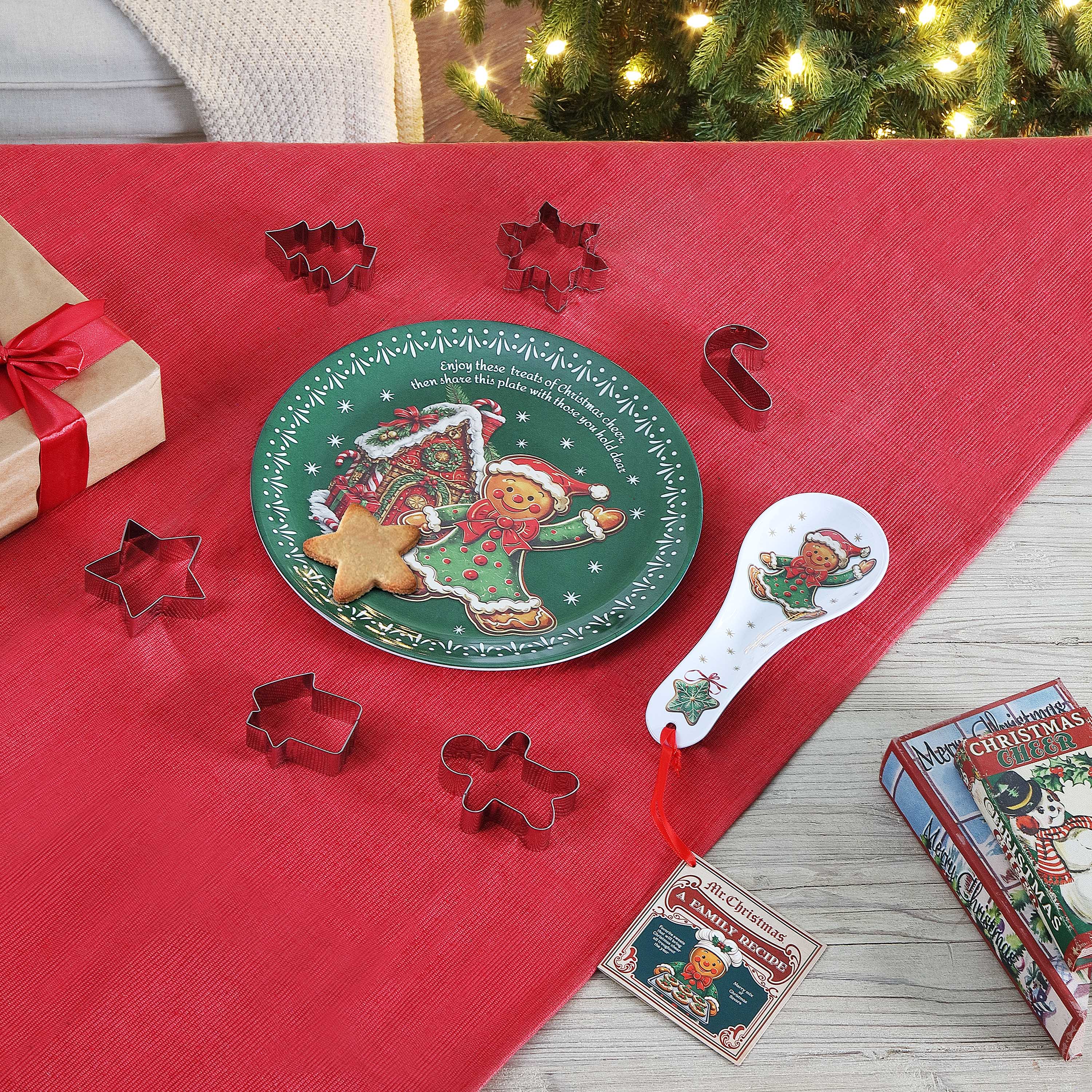 A Christmas-themed cookie cutter set arranged on a red table runner, with a green decorative plate, metal cookie cutters in star, tree, and gingerbread shapes, a white spoon, and a gift box in the background near a lit Christmas tree.