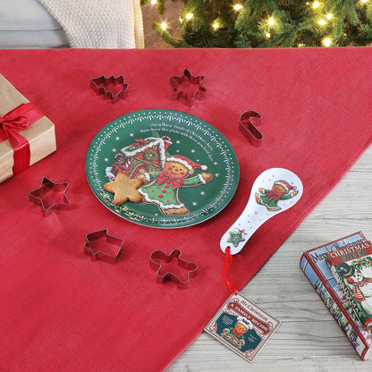 A Christmas-themed cookie cutter set arranged on a red table runner, with a green decorative plate, metal cookie cutters in star, tree, and gingerbread shapes, a white spoon, and a gift box in the background near a lit Christmas tree.