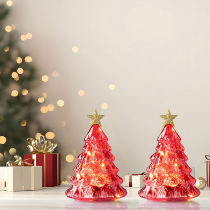 Two red Christmas tree-shaped table lamps with warm white lights and gold star toppers, displayed on a tabletop with wrapped gifts and blurred holiday lights in the background.