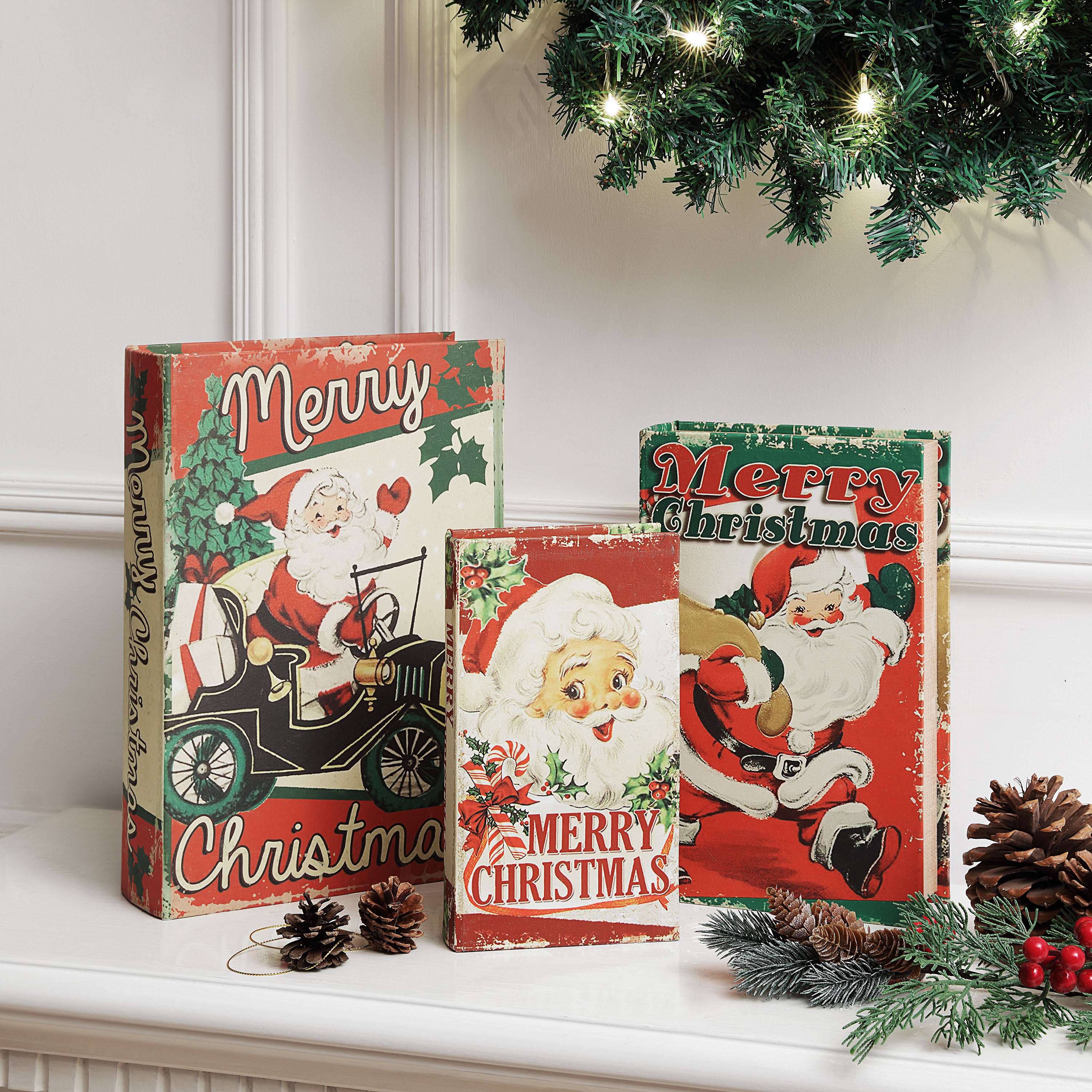 Three vintage-style Christmas book covers or decorative book boxes displayed on a mantel, featuring Santa Claus illustrations and Merry Christmas text, with pinecones, evergreen branches, and a Christmas tree branch in a holiday setting.