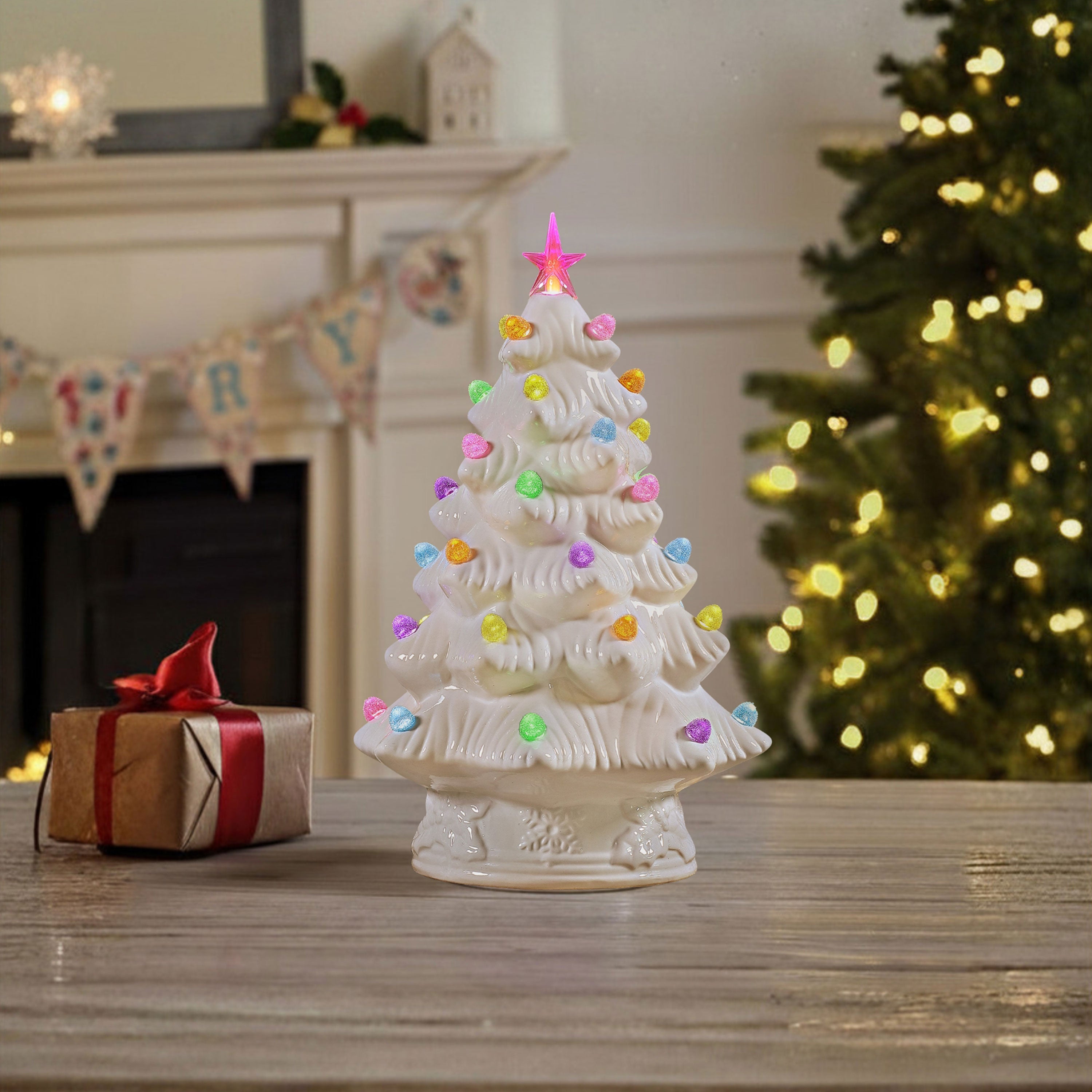 A white ceramic Christmas tree ornament with colorful glowing gumdrop lights and a pink star topper, displayed on a wooden table next to a wrapped gift box, with a fireplace and a lit Christmas tree in the background.